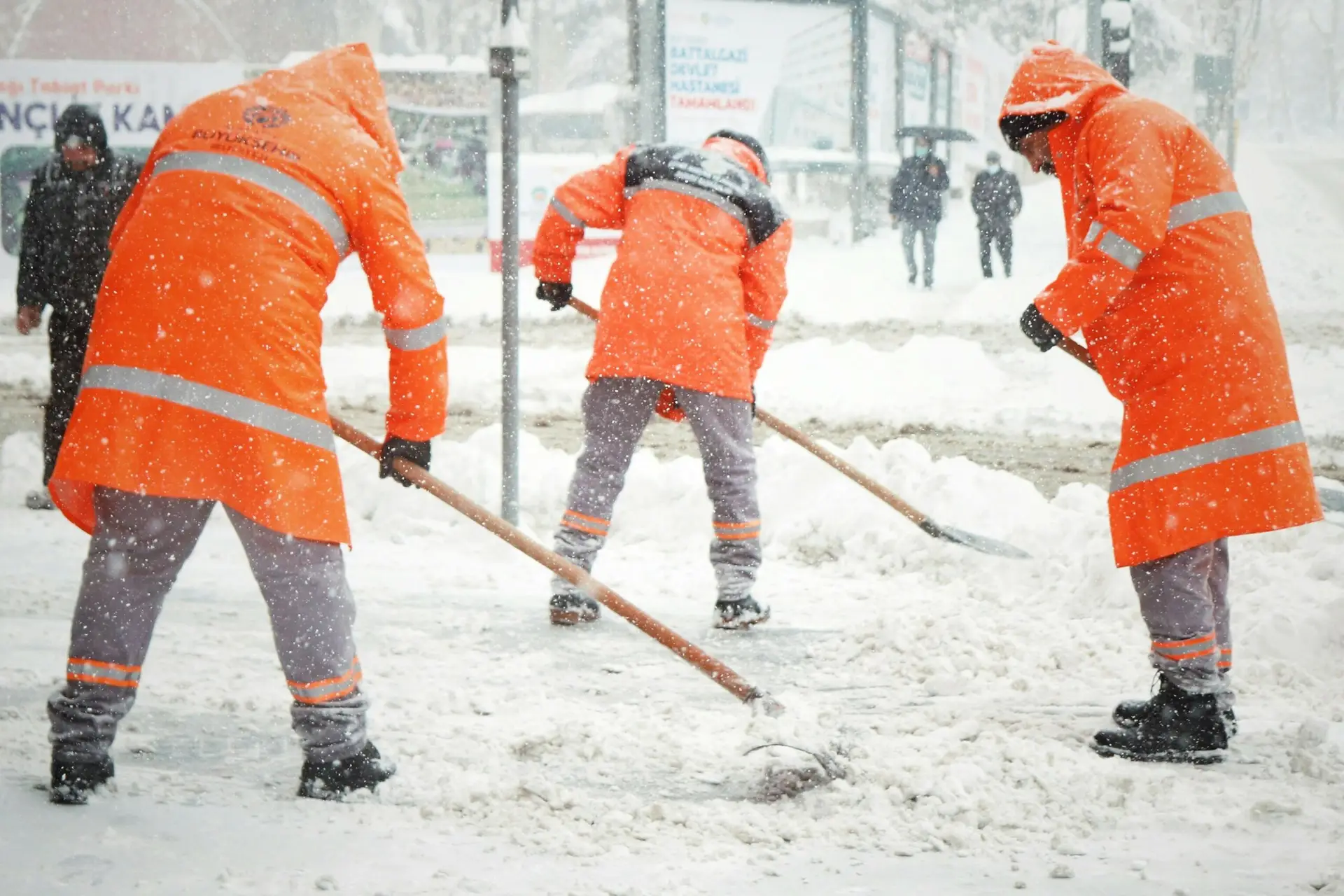 Trois agents municipaux en manteaux orange déneigent une rue.