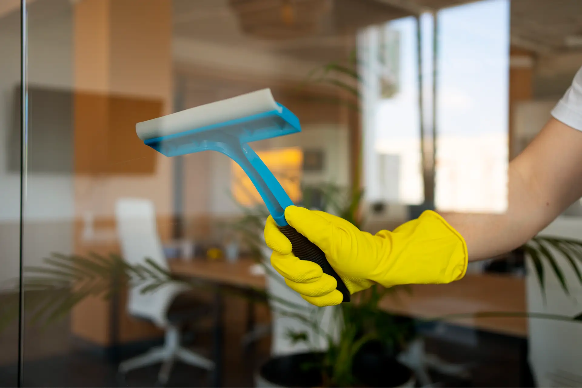 Main gantée de jaune nettoyant une vitre avec une raclette.
